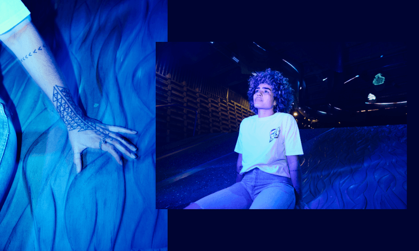 A person with curly hair and a geometric tattoo on their arm sits on a textured, wave-like surface under blue and purple lighting, looking thoughtfully into the distance. A close-up of their tattooed hand is shown on the left.