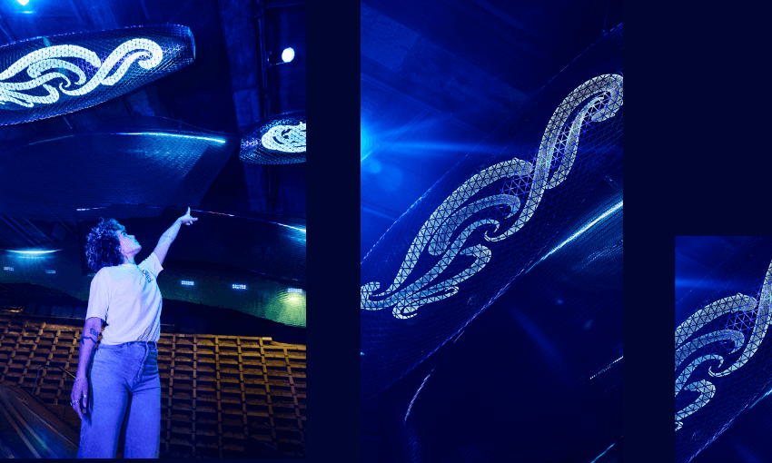 A person standing under large, illuminated, wave-shaped sculptures with intricate patterns, reaching upward in a space lit with vibrant blue lighting. Rows of empty seats are visible in the background.