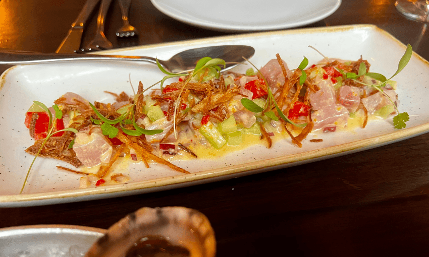 A rectangular plate with a ceviche dish featuring diced raw fish, thinly sliced fried onions, red peppers, greens, and a light yellow sauce, served on a dark wooden table with a spoon and cutlery in the background.