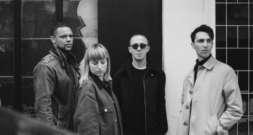 A black and white photo of a band: three men and one woman. They all have jackets on and look aloof.