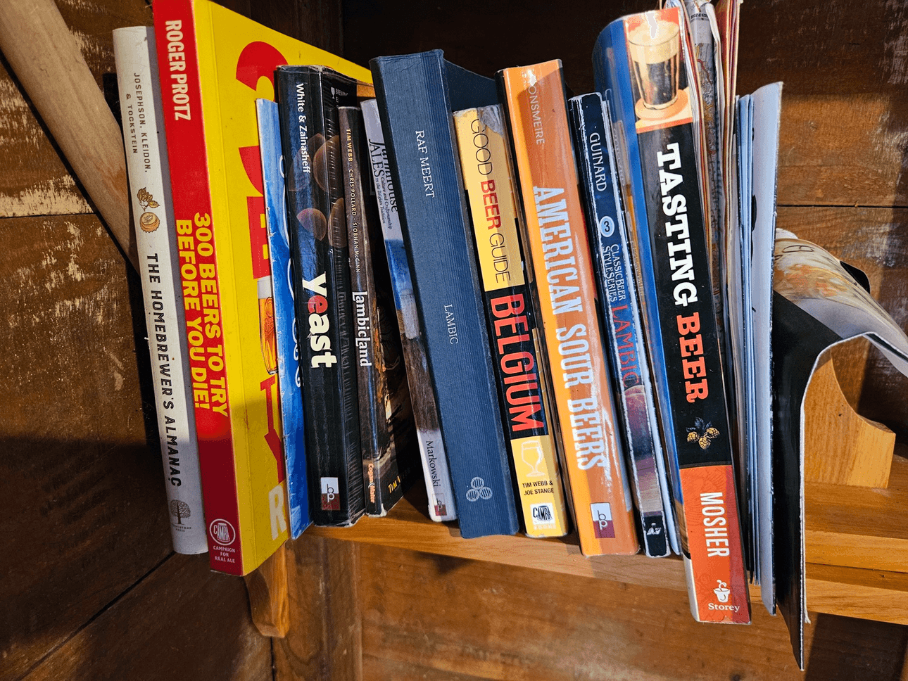 A collection of books about beer and brewing, including titles such as "yeast," "Beer Guide Belgium," "American Sour Beers," and "Tasting Beer," arranged on a wooden shelf.