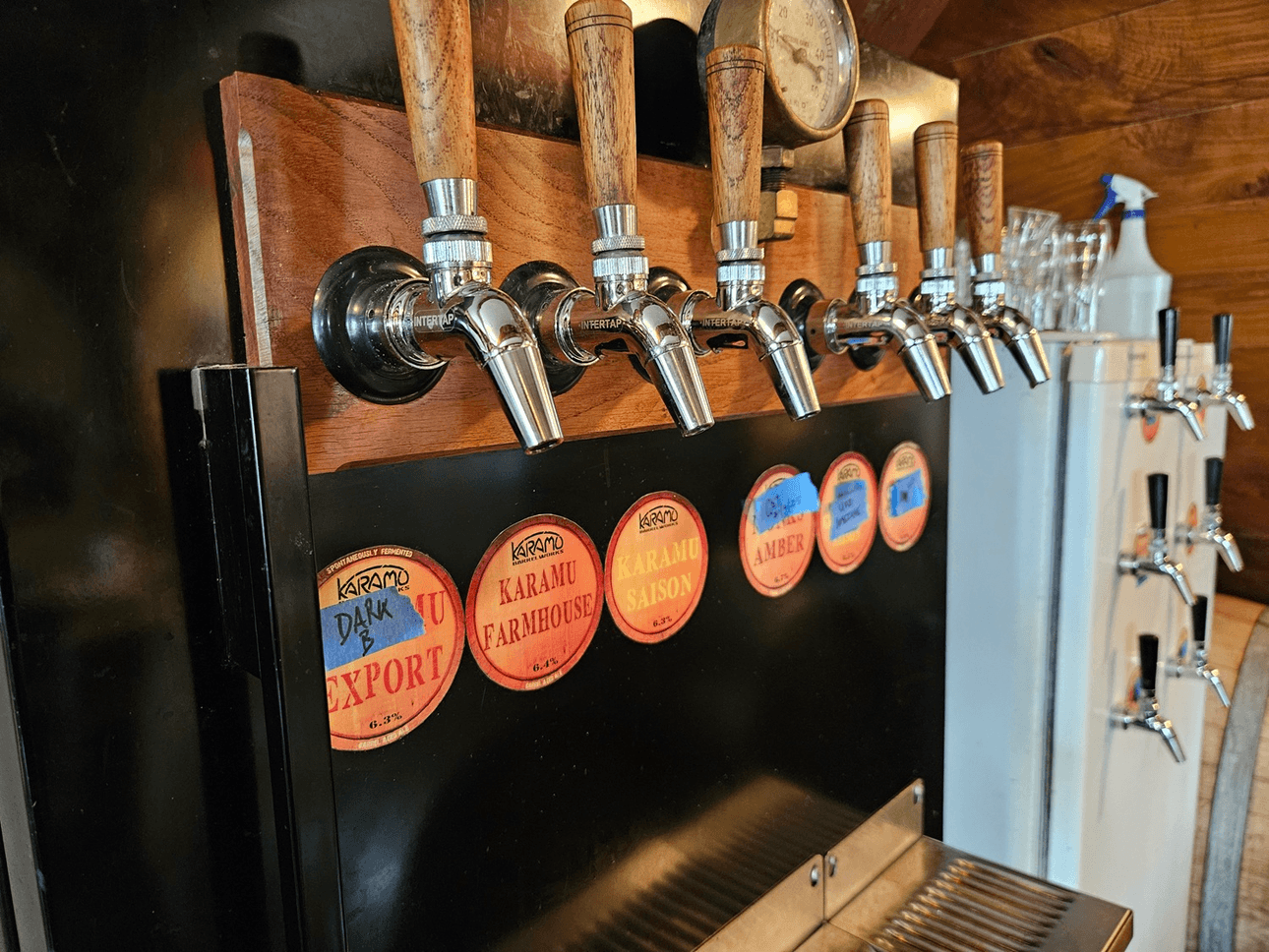A close-up of six beer taps with wooden handles mounted on a black bar front, each tap labeled with different beer names like "Karamu Dark Export" and "Karamu Farmhouse." Glassware and a sanitizer bottle are visible in the background.