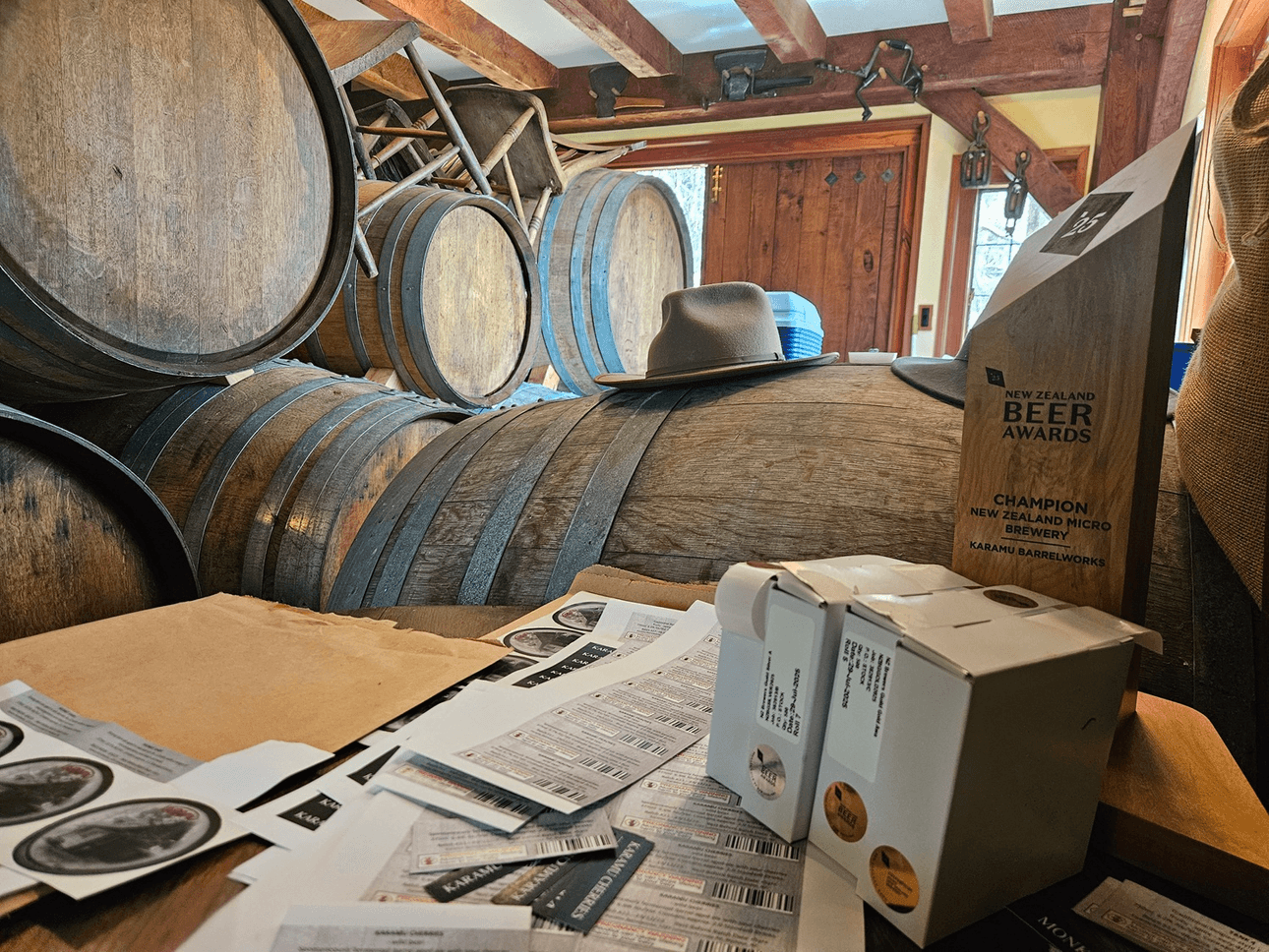 A rustic room with large wooden barrels, scattered papers, labels, a beer award trophy, and a light grey hat resting on one of the barrels. Sunlight streams in through a window, highlighting the cozy atmosphere.