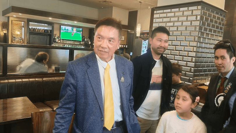 A man in a blue jacket and yellow tie stands in a restaurant with three other people, including a young girl. Behind them, a TV shows a sports game. The setting appears casual and lively.