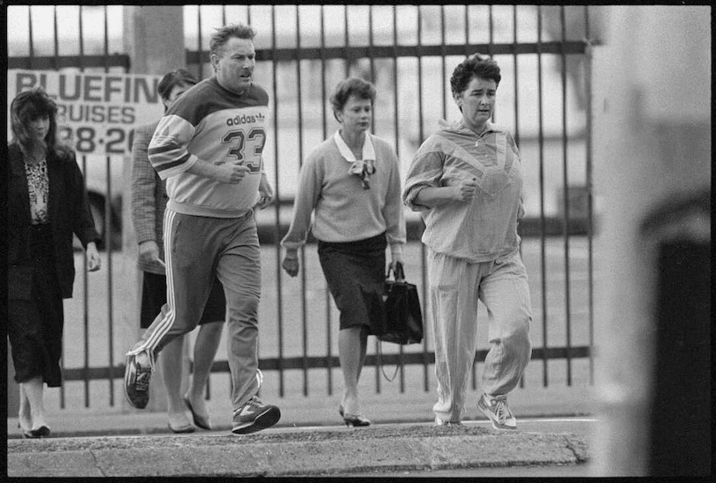 Four adults cross a city street; two wear business attire, while a man and woman in tracksuits jog beside them. A fence and billboard are visible in the background. The image is in black and white.