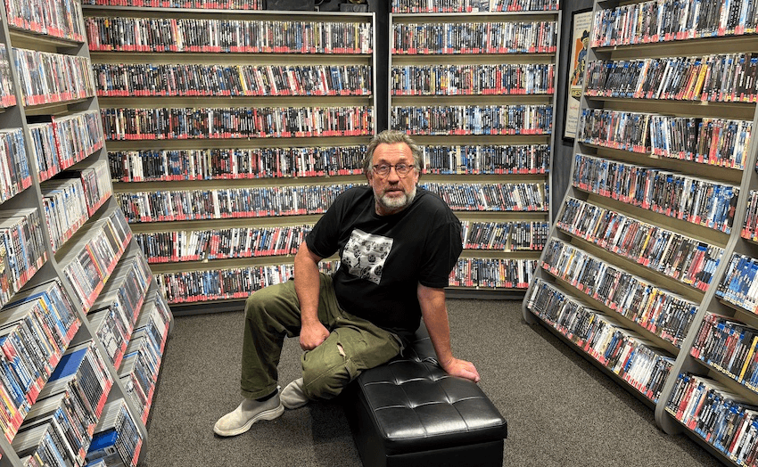 A man sits in the middle of a room surrounded by hundreds of DVDs