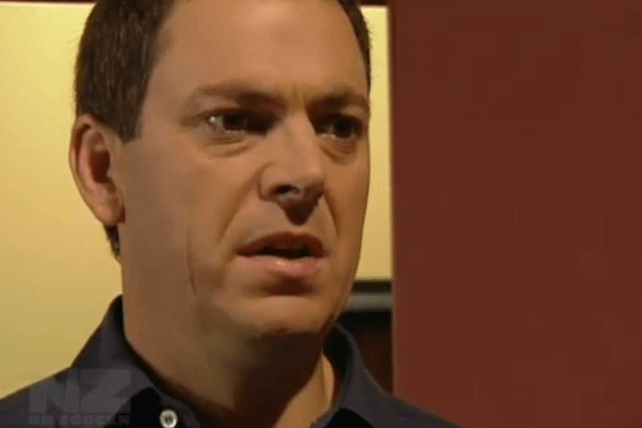 A man with short brown hair and a concerned expression is standing indoors against a yellow and brown background. He is wearing a dark collared shirt.