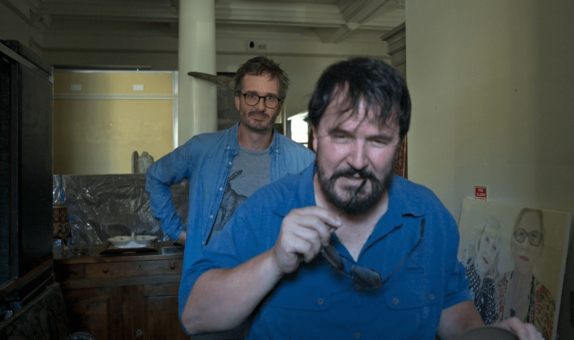 Two men stand indoors. The man in front, with dark hair and beard, holds sunglasses and looks toward the camera. The man behind him, with glasses and a beard, stands with his hand on his hip. Paintings are visible on the right.