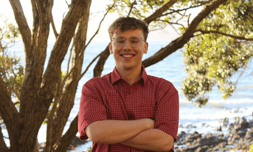 A young man in glasses and a red plaid shirt stands smiling with his arms folded, in front of a tree and beach.