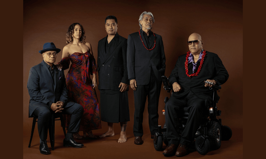 Five people pose against a brown background, dressed in formal and traditional attire. One man sits in a wheelchair, another on a stool, while the others stand, including a woman in a red patterned dress.