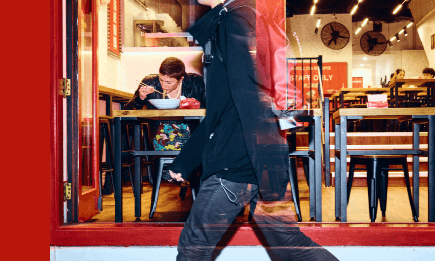 A person wearing black walks past a restaurant window. Inside, another person sits alone at a table, eating from a bowl. The interior is brightly lit with red and wood décor and empty tables.
