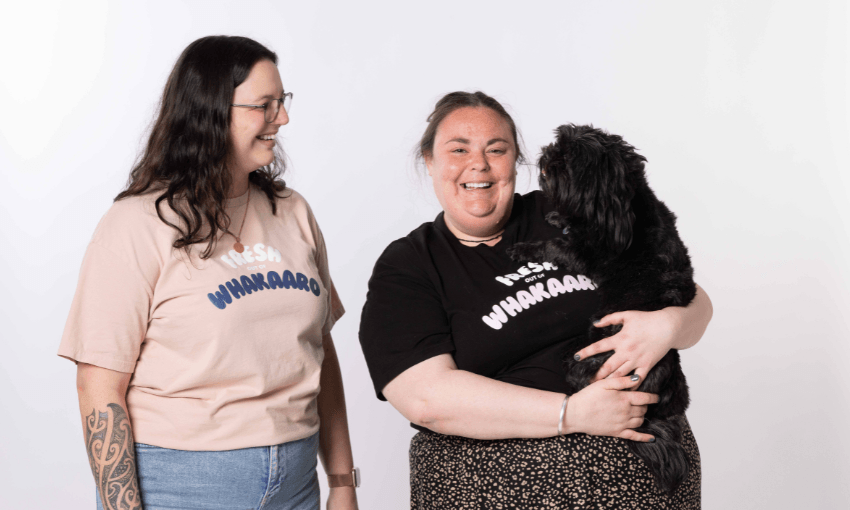 Two women smile together. One wears glasses and a beige shirt; the other, in a black shirt, holds a small black dog. Both shirts have the word “WHAKAARO” printed on them. The background is plain white.