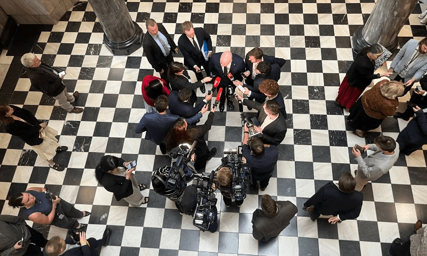 A bird's eye view shot of Christopher Luxon being interviewed by media on parliament's tiles.