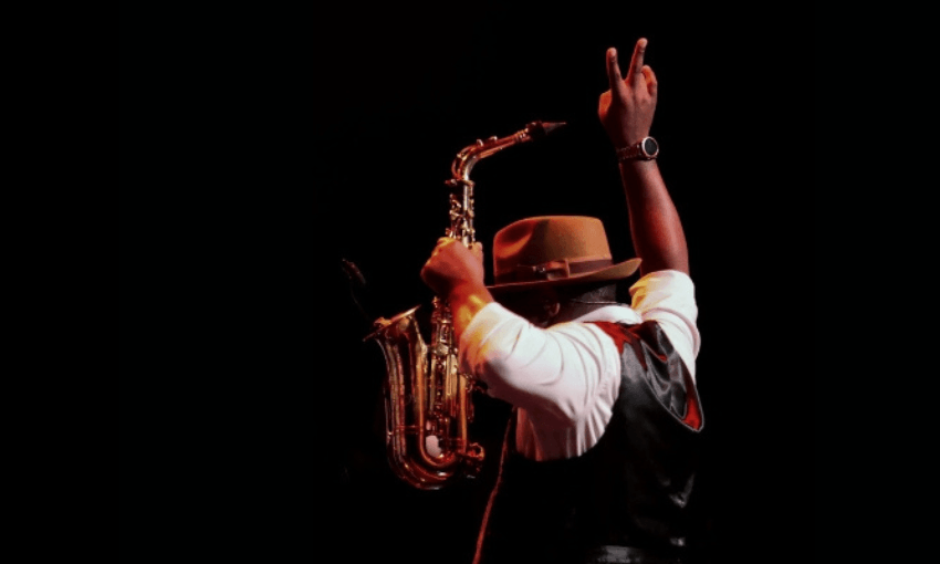 A musician in a hat holds a saxophone under one arm and raises his other hand making a peace sign, standing against a dark background with dramatic lighting.