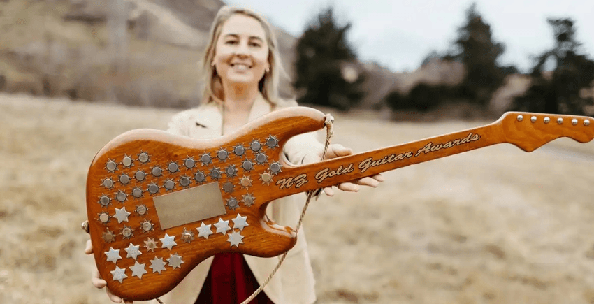 A woman standing outside in a brown grassy field, smiling, and holding a guitar bedazzled with gold coins, for us to see.