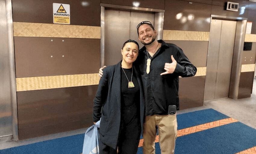 Two people stand together smiling in front of elevators. One person has an arm around the other and is making a shaka sign with their hand. Both are dressed in dark clothing and wear green pendants. A warning sign is visible above them.