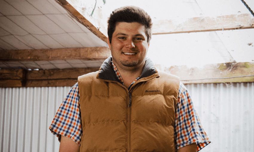 A man with short dark hair smiles while wearing a tan puffer vest over a red, white, and blue plaid shirt. He stands indoors beneath a partially open roof with exposed wooden beams and corrugated metal walls.