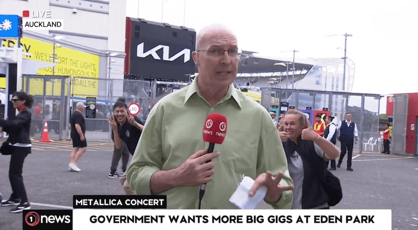 TVNZ reporter Simon Mercep talks to camera as Metallica fans in the background wave to the camera