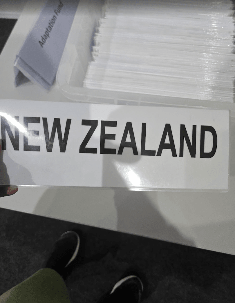 A person is holding a sign that reads "NEW ZEALAND" in bold black letters. In the background, there are labeled folders, one of which reads "Adaptation Fund.