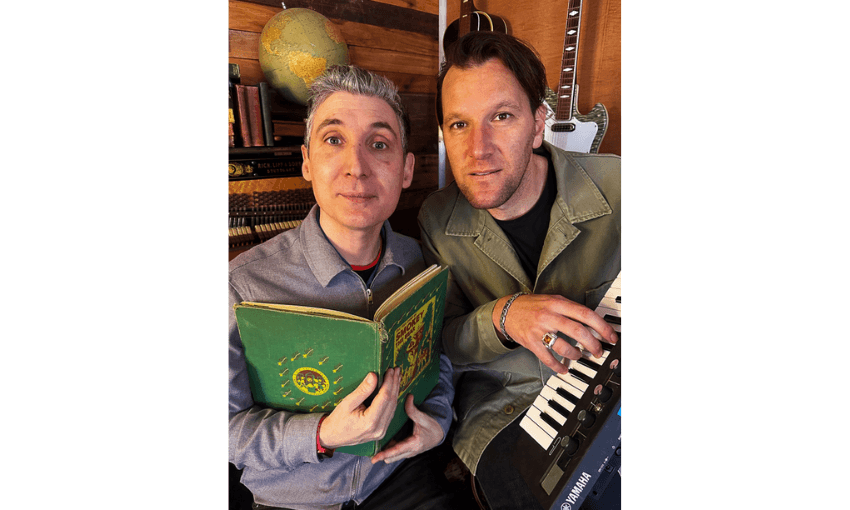 A photo of two men, one holding an open book and the other playing on a keyboard.