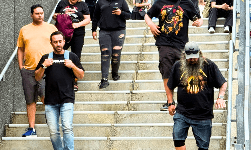 A group of people, casually dressed, walk down outdoor concrete stairs off teh train on their way to Metallica in Auckland. Some are wearing graphic t-shirts and jeans; one man has a long gray beard. Most appear to be looking at their phones or straight ahead.