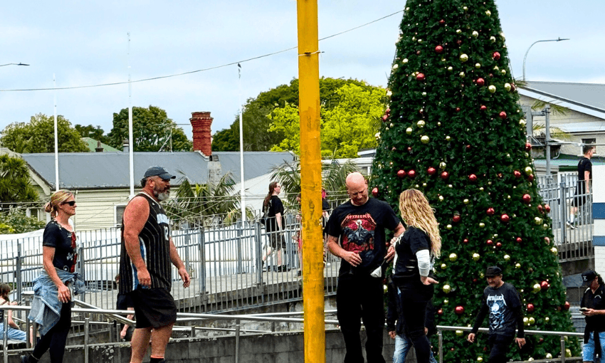 Metallica fans walk near a large outdoor Christmas tree decorated with red and gold ornaments. Some are on ramps and steps, dressed casually, with buildings and greenery visible in the background.