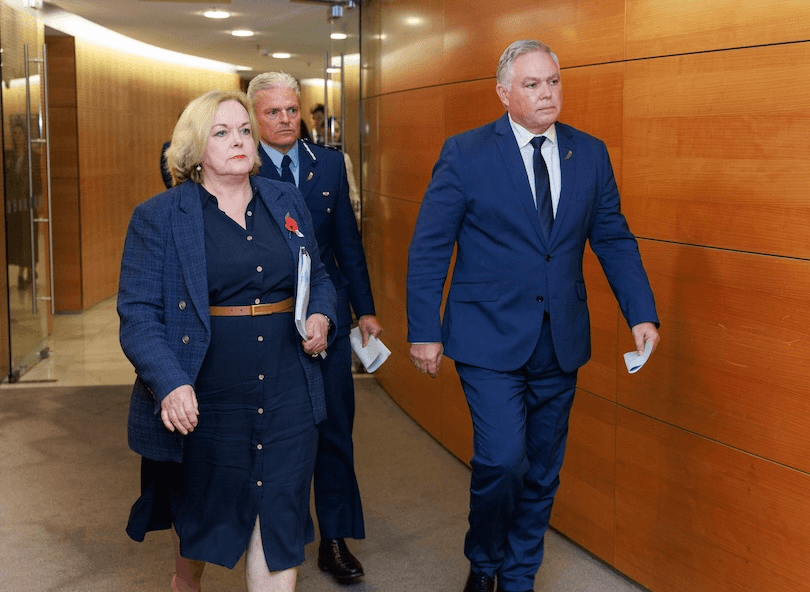 Three people in formal attire walk down a hallway with wooden panel walls. The woman in front wears a dark dress and blazer, followed by two men in blue suits, one in uniform. They appear focused and serious.