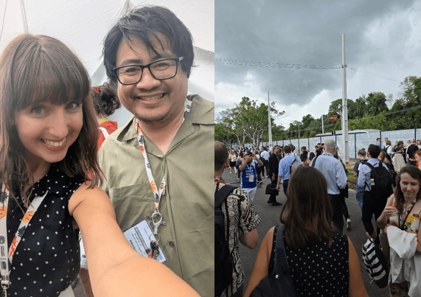 A woman and a man smile for a selfie at an outdoor event. On the right, a crowd of people, some wearing lanyards, walk along a street under a cloudy sky lined with trees and utility poles.