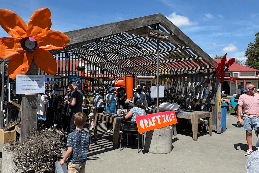 An outdoor marae-like structure filled with people beneath signs reading "Kotahitanga" and "Craft Zone" 