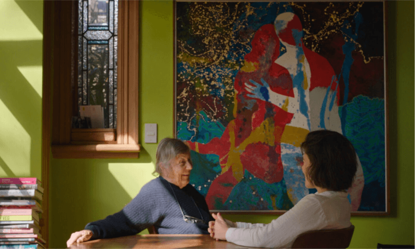 an older woman speaks to a younger woman at a table. a colourful large painting hangs on the green wall behind them