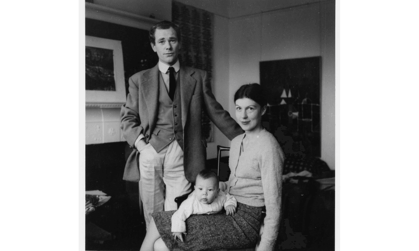a black and white family portrait taken inside a home. A man stands in a three piece suit and a woman sits wearing a cardigan and a long skirt. Their baby lies on the woman's lap