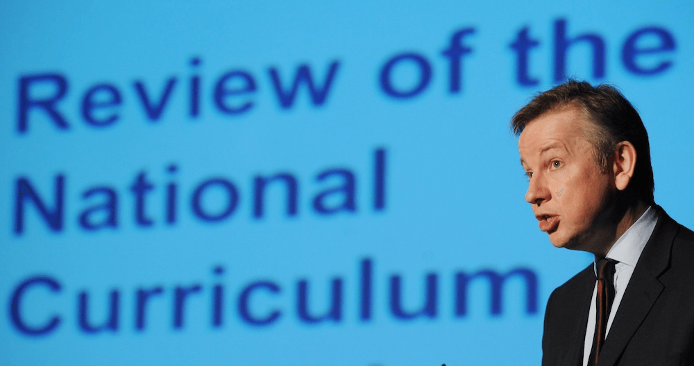 A man in a suit speaks in front of a blue screen displaying large text that reads “Review of the National Curriculum.”.