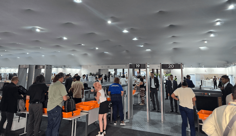 People line up for security screening at a busy indoor checkpoint with metal detectors and security personnel. Some are placing belongings in orange trays on conveyor belts. The ceiling has a unique, wavy pattern.