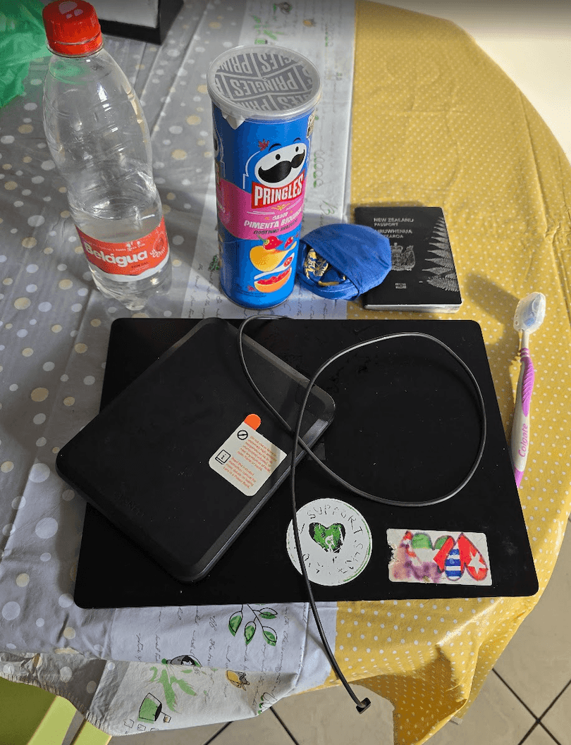 A table with a bottle of water, a Pringles can, a passport, a face mask, a toothbrush, a cable, and two black electronic devices, one with stickers, all on a patterned tablecloth.