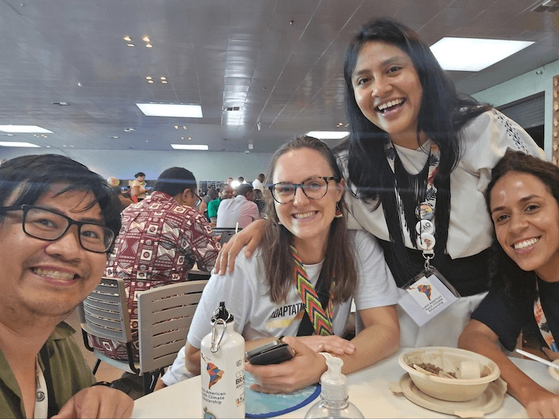 Four people smiling at a cafeteria table, three seated and one standing behind them, with food, drinks, and personal items on the table. Other diners are visible in the busy background.