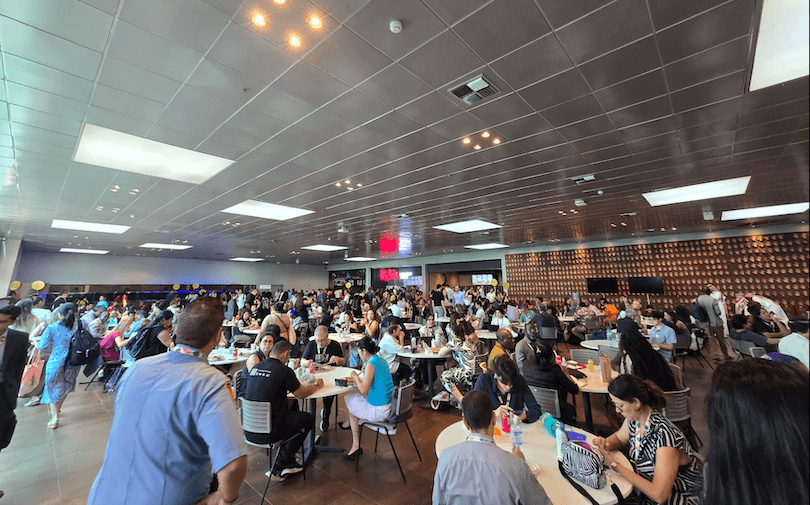 A large, crowded cafeteria filled with people sitting at round tables, eating, talking, and socializing under bright ceiling lights with a modern interior design.