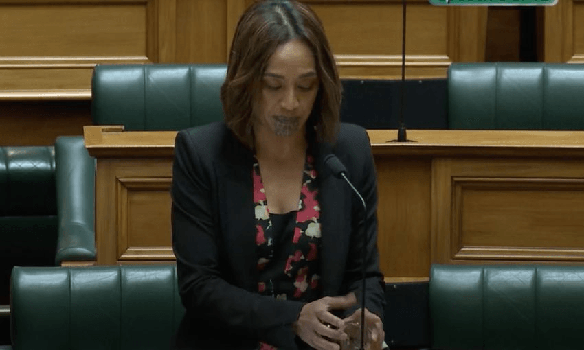Oriini Kaipara stands behind in her bench in the House, catching a glass of water in her hands as it falls over.