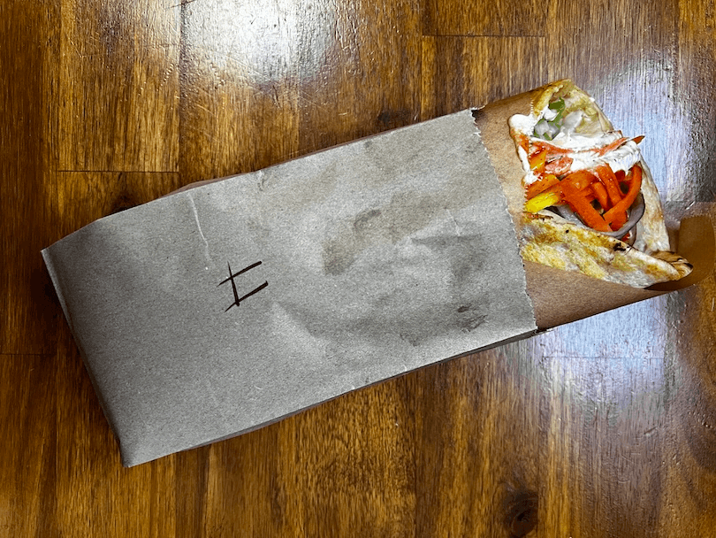 kebab or pita with visible fillings like lettuce, tomatoes, and sauce peeks out from a brown paper wrapper, resting on a wooden table.