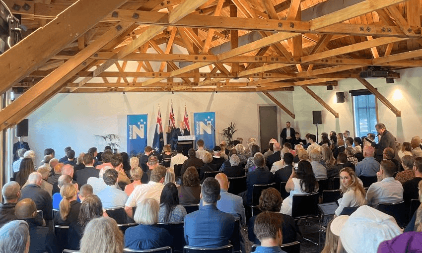 Christopher Luxon stands at a podium with National Party banners behind him, and a crowd of seated supporters in front.