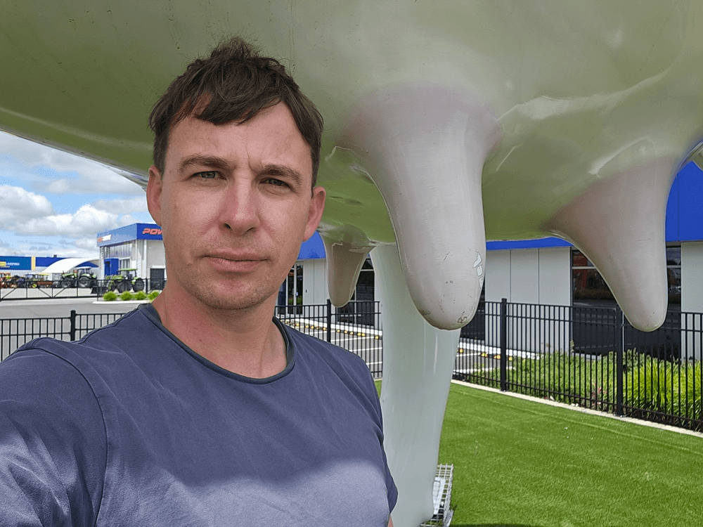 A man in a grey shirt takes a selfie in front of a large statue of a cow's udder outdoors, with buildings, a black fence, and green grass in the background.