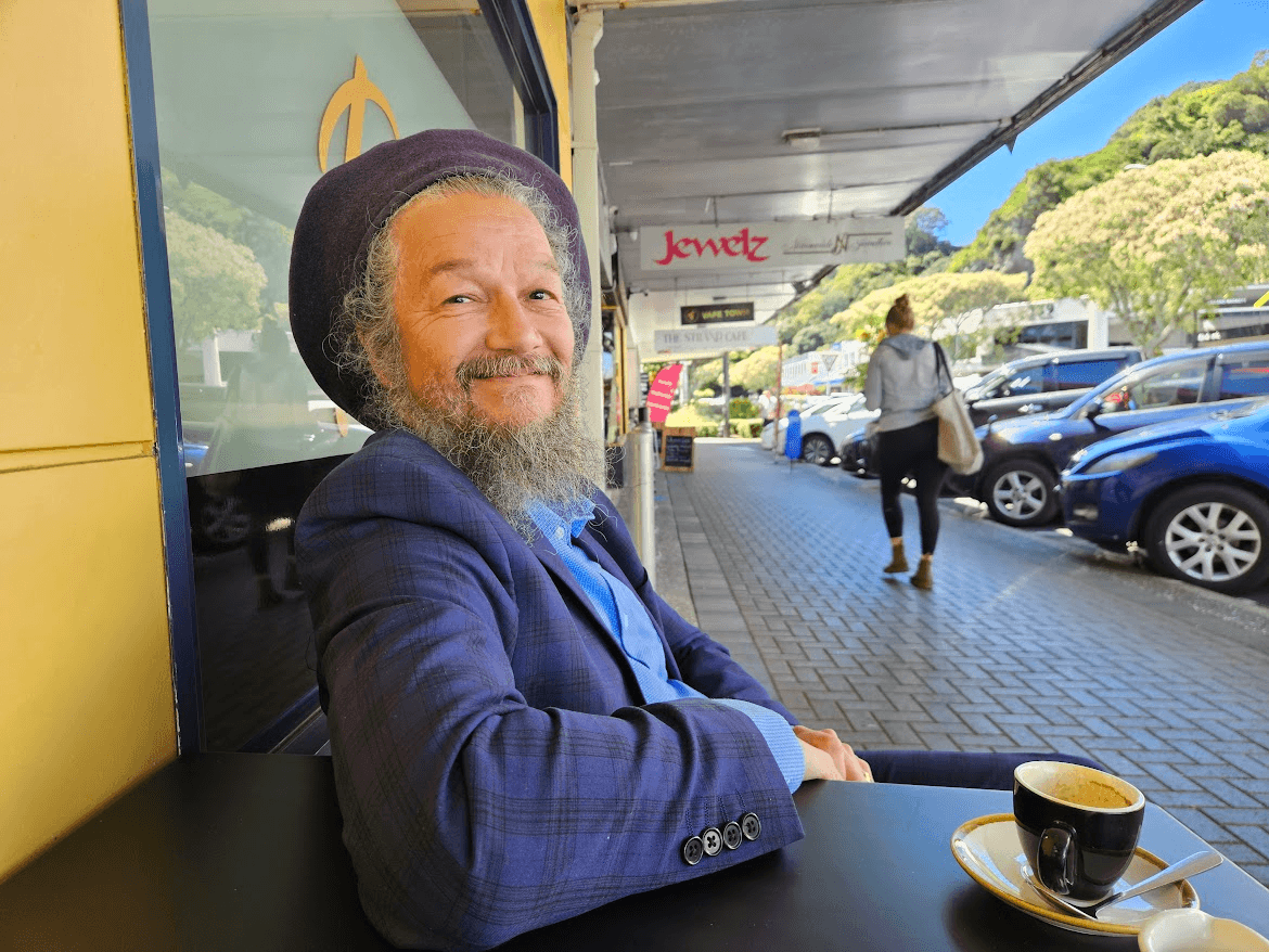 A smiling man with a gray beard and a hat sits at an outdoor café table with a coffee cup, on a sunny street with cars parked and people walking by.