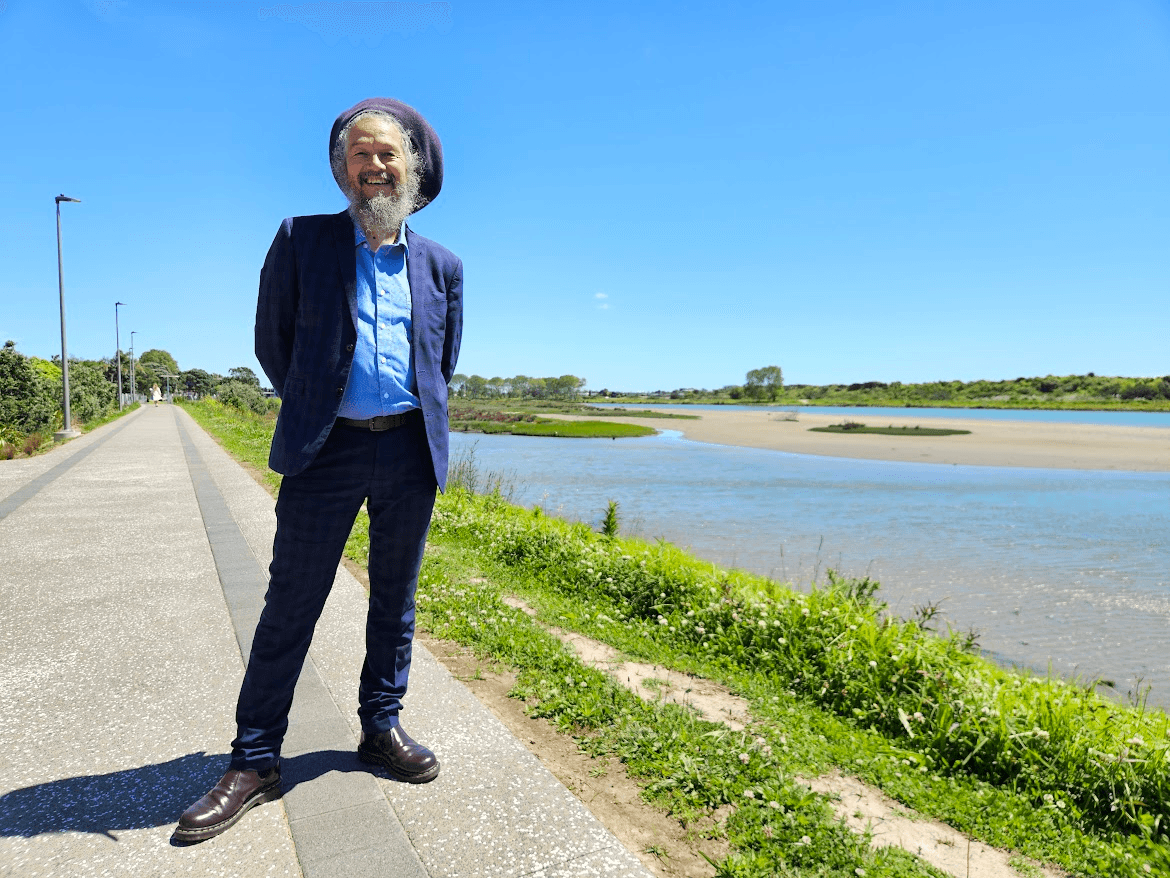 A smiling man with a beard and hat stands on a paved path beside a river or estuary, dressed in a blue suit and boots, with greenery and a clear blue sky in the background.