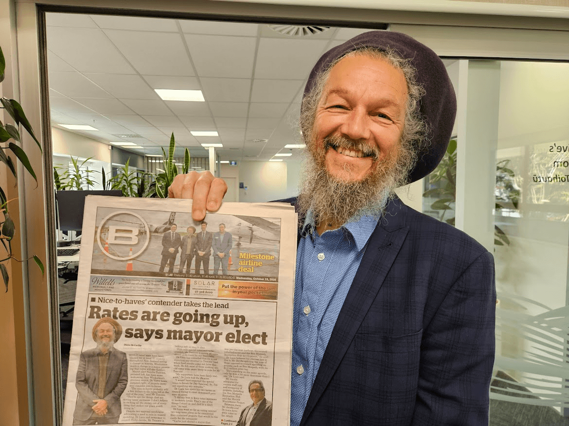 A smiling man with a gray beard and dreadlocks, wearing a dark blazer and hat, holds up a newspaper featuring his photo on the front page with the headline "Rates are going up, says mayor elect.