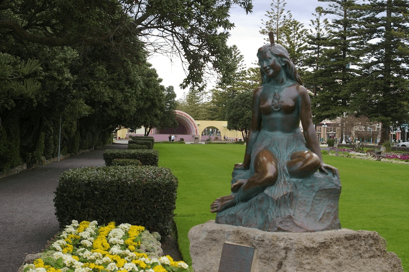 A bronze statue of a girl sits on a rock in a garden, surrounded by neatly trimmed bushes and yellow flowers. In the background, there’s a pink bandstand and tall trees lining a grassy park.