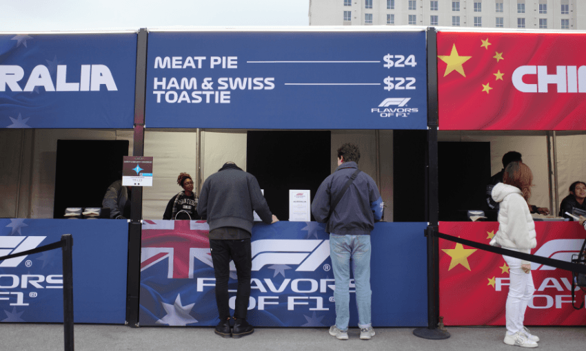 two men wearing jackets and long pants face away from the camera and stand at a blue food stall. above them in white writing is the menu "meat pie $24, ham and swiss toastie $22"