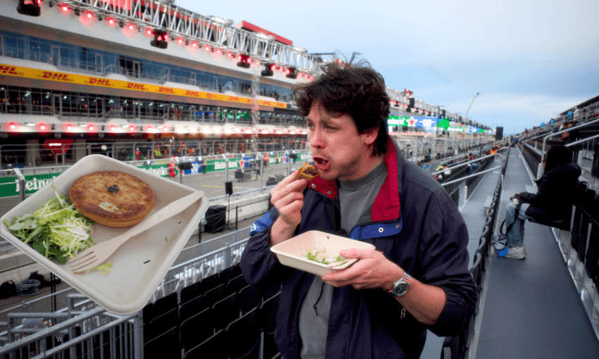 A man wearing a jacket stands in the stands of a grand prix track and is mid-bit of a pie