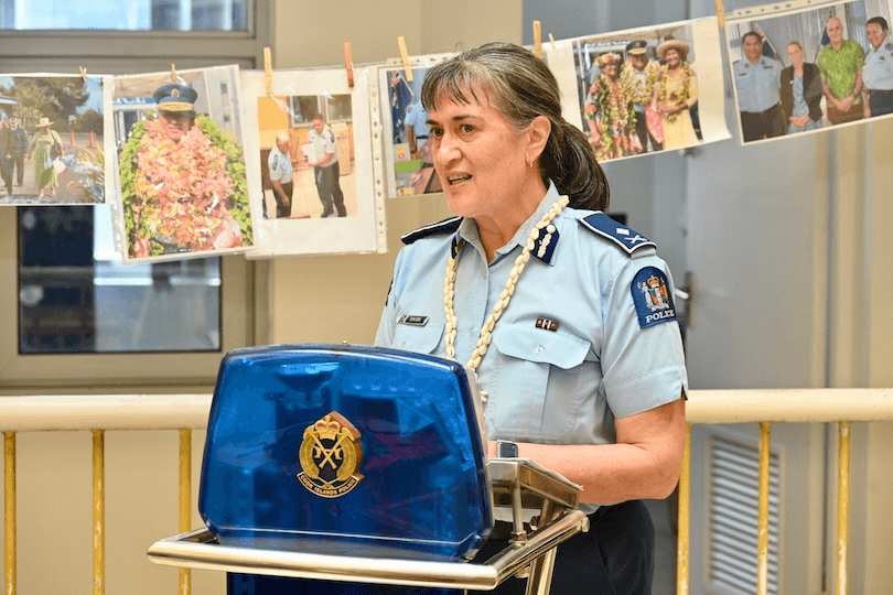 A policewoman in uniform speaks at a podium. Behind her, photos of police officers and events are displayed on a string. She wears a white lei and stands indoors, smiling as she addresses an audience.