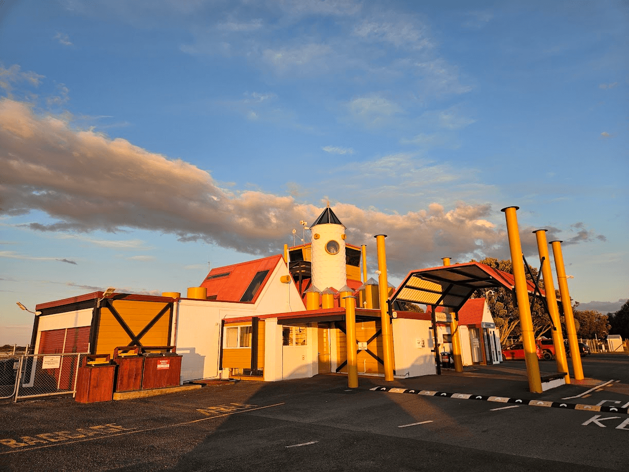 A quirky building with yellow pillars, red roofs, and a white tower with a blue peaked roof, lit by golden sunlight. A dramatic cloud stretches across the sky above, and a red car is parked nearby.