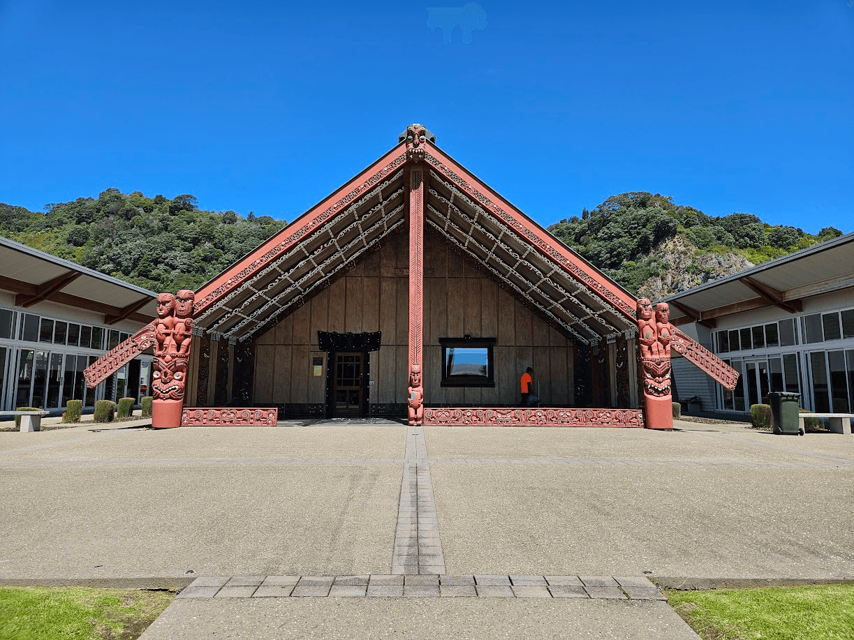 A traditional Māori meeting house with ornate carvings under a clear blue sky, surrounded by modern buildings and greenery, with a person in an orange shirt near the entrance.