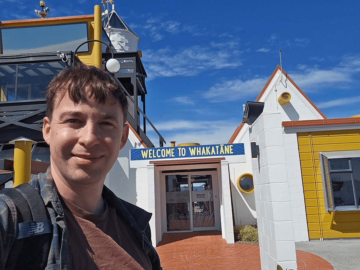 A man with brown hair smiles at the camera in front of a building with a sign that reads “Welcome to Whakatāne” against a blue sky.